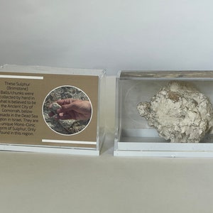 Könnte beinhalten: Zwei transparente Vitrinen mit weißen Sockeln. Die Vitrine links hat ein braunes Etikett mit Text über Schwefel (Schwefel) Kugeln/Brocken, die von Hand in der antiken Stadt Gomorrha gesammelt wurden, unterhalb von Masada in der Region des Toten Meeres in Israel. Die Vitrine rechts enthält ein großes, weißes, steinartiges Stück Schwefel.
