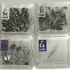 May include: A collection of small, clear plastic containers, each containing a sample of a different element. The elements are labelled with their atomic number and symbol, and some have their names written below. The elements include aluminium, sulfur, scandium, titanium, cobalt, germanium, rhodium, indium, antimony, caesium, lutetium, and osmium.
