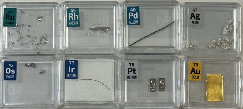 May include: A collection of small, clear plastic containers, each containing a sample of a different element. The elements are labeled with their atomic number and symbol, and their names are listed below. The elements included are Ruthenium, Rhodium, Palladium, Silver, Osmium, Iridium, Platinum, and Gold.