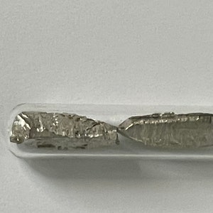 Puede incluir: Dos piezas de metal de color plateado se encuentran dentro de un tubo de vidrio transparente. Las piezas de metal tienen una forma irregular y un acabado brillante y metálico.