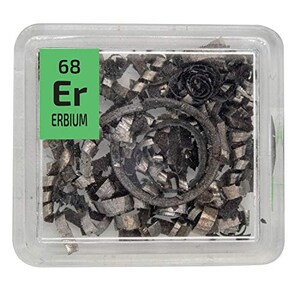 Puede incluir: Un recipiente de plástico transparente que contiene virutas de erbio metálico. Una etiqueta verde muestra "68 Er ERBIO". Las virutas son plateadas y grises, con un objeto oscuro y circular en el centro.