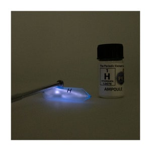 May include: A small glass ampoule containing hydrogen gas. The ampoule is labelled with the periodic table element symbol for hydrogen, "H", and the atomic number "1". The ampoule is sitting on a white surface next to a clear glass slide with the letter "H" written on it. The slide is glowing blue.