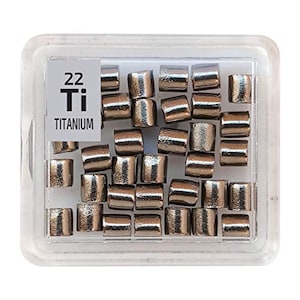 Puede incluir: Un recipiente de plástico transparente lleno de pequeñas pastillas de titanio de color plateado. El recipiente tiene una etiqueta que dice "22 Ti TITANIUM".