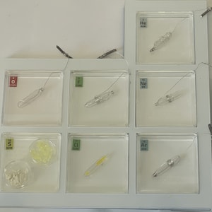 Puede incluir: Una vitrina blanca con nueve recipientes de vidrio transparente, cada uno con un elemento diferente. Los elementos están etiquetados con su número atómico y su nombre. Los elementos son Helio, Oxígeno, Flúor, Neón, Azufre, Cloro, Argón y una muestra de azufre.