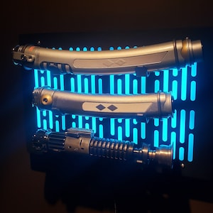 May include: Three lightsabers mounted on a black wall display with a blue light grid pattern. The lightsabers are silver and black with a gold accent.
