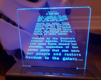 May include: A blue and white illuminated sign with the text "Episode IV A NEW HOPE" and a description of the plot of the movie Star Wars: A New Hope. The sign is on a black base.