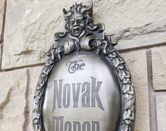 May include: A silver-colored metal sign with a decorative frame and a gargoyle at the top. The sign reads "The Novak Manor".