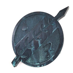 May include: A dark green and blue circular metal Mandalorian symbol with a spear through it. The symbol has a stylized water drop design and a crescent moon pattern.