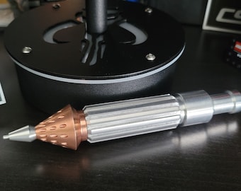 May include: A silver and copper-colored tool with a pointed tip and a textured, ribbed body. The tool is lying on a dark surface.