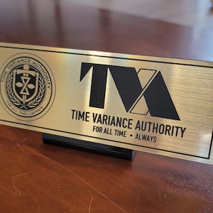以下が含まれることがあります： 「Time Variance Authority」と「TVA」のイニシャルが黒で書かれた金色の金属製の銘板。銘板には、剣、砂時計、盾が描かれた円形のロゴも含まれています。