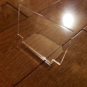 May include: A clear acrylic phone stand with a simple design. The stand is angled to hold a phone upright.