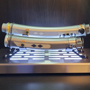 May include: Two silver lightsaber hilts with black accents are displayed on a metal stand with a grid pattern. The stand has a white light shining through the grid.