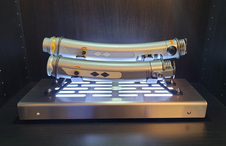May include: Two silver lightsaber hilts with black accents are displayed on a metal stand with a grid pattern. The stand has a white light shining through the grid.