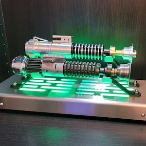 May include: Two silver and black lightsabers with green lights on a silver metal stand with a green light grid.