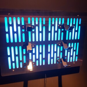 May include: A metal wall art piece with a blue light behind it. The art piece is shaped like a grid with a pattern of lines and holes. There are several metal pieces attached to the front of the art piece.