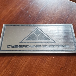 May include: A rectangular, brushed metal plaque with the words "CYBERDYNE SYSTEMS" in a sans-serif font. Above the text is a triangular graphic with a geometric design. The plaque has a silver finish.