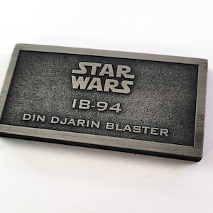 Może przedstawiać: Srebrna metalowa tabliczka z tekstem "STAR WARS" w dużych literach, "IB-94" w dużych literach i "DIN DJARIN BLASTER" w dużych literach.