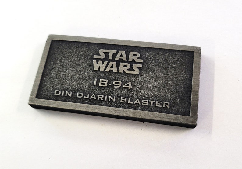 Może przedstawiać: Srebrna metalowa tabliczka z tekstem "STAR WARS" w dużych literach, "IB-94" w dużych literach i "DIN DJARIN BLASTER" w dużych literach.