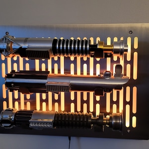 May include: Three silver lightsabers mounted on a metal wall panel with a grid pattern. The lightsabers are illuminated from behind, creating a warm glow.