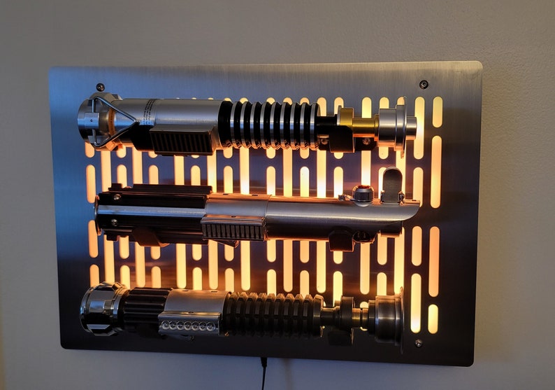 May include: Three silver lightsabers mounted on a metal wall panel with a grid pattern. The lightsabers are illuminated from behind, creating a warm glow.