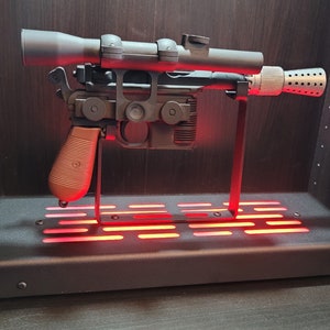 May include: A black and silver blaster pistol with a brown handle and a scope on top, displayed on a black stand with red lights.