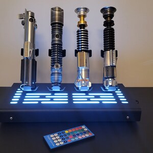 Pode incluir: Quatro r&eacute;plicas de sabres de luz em um suporte preto com luz azul. O suporte tem um controle remoto com v&aacute;rios bot&otilde;es coloridos.