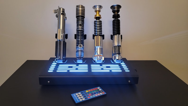 Pode incluir: Quatro r&eacute;plicas de sabres de luz em um suporte preto com luz azul. O suporte tem um controle remoto com v&aacute;rios bot&otilde;es coloridos.