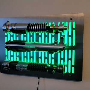 May include: A stainless steel wall mount with a green light grid pattern. Three silver lightsabers are mounted on the wall mount. The lightsabers are lit up with a green glow.