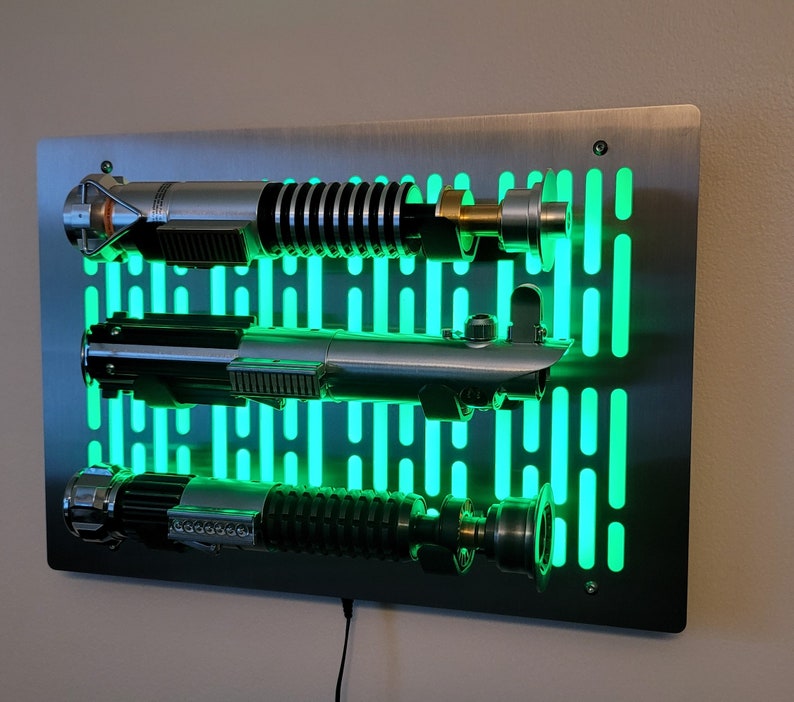 May include: A stainless steel wall mount with a green light grid pattern. Three silver lightsabers are mounted on the wall mount. The lightsabers are lit up with a green glow.