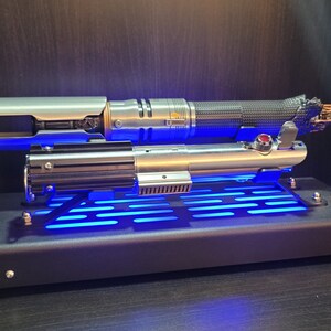 May include: A silver and black lightsaber with a blue light display on a black stand. The lightsaber is on a black stand with a blue light display.