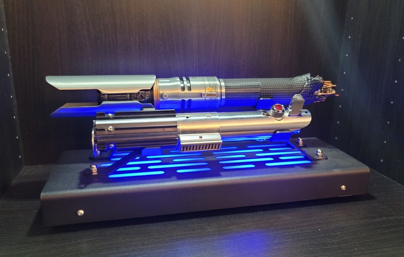 May include: A silver and black lightsaber with a blue light display on a black stand. The lightsaber is on a black stand with a blue light display.