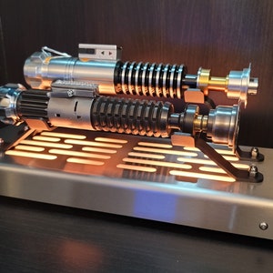 May include: A silver and black lightsaber replica with a light-up base. The lightsaber is displayed on a metal stand with a grid pattern.