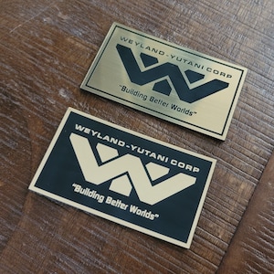 Op de afbeelding: Twee Weyland-Yutani Corp. logo's. Het bovenste logo is goud met een zwarte "W" en het onderste logo is zwart met een gouden "W". Beide logo's hebben de tekst "Building Better Worlds" eronder.