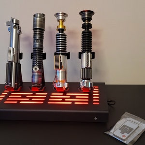 Pode incluir: Quatro r&eacute;plicas de sabres de luz em um suporte preto com luzes LED vermelhas. Os sabres de luz s&atilde;o prateados, pretos, dourados e uma combina&ccedil;&atilde;o de prata e preto.