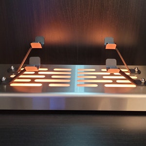 May include: A silver metal platform with a grid pattern and two black metal arms with copper-colored accents. The platform is illuminated from below, creating a warm glow.