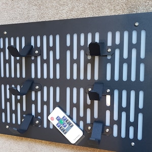 May include: A black metal wall organizer with a white grid pattern and multiple hooks. The organizer has a Star Wars inspired design. A remote control is sitting on the organizer.