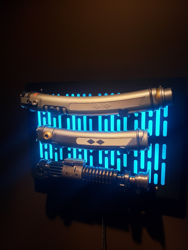May include: Three lightsabers mounted on a black wall display with a blue light grid pattern. The lightsabers are silver and black with a gold accent.