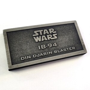 Może przedstawiać: Srebrna metalowa tabliczka z tekstem "STAR WARS" w dużych literach, po kt&oacute;rym następuje "IB-94" i "DIN DJARIN BLASTER" w dużych literach.
