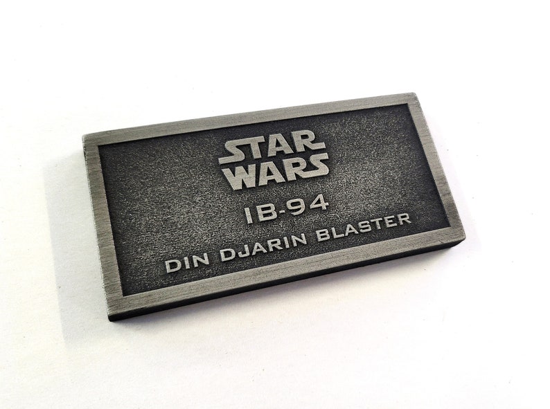 Może przedstawiać: Srebrna metalowa tabliczka z tekstem "STAR WARS" w dużych literach, po kt&oacute;rym następuje "IB-94" i "DIN DJARIN BLASTER" w dużych literach.