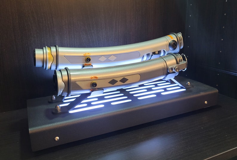 May include: Two silver lightsaber hilts with black accents and a diamond-shaped design on the side are displayed on a black stand with a white light grid.
