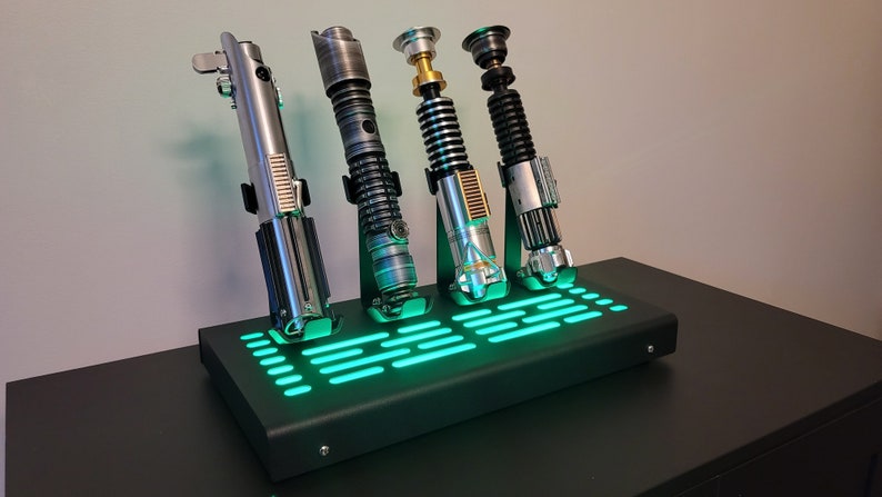 Pode incluir: Quatro r&eacute;plicas de sabres de luz em um suporte preto com luz verde. O suporte tem um padr&atilde;o de grade e os sabres de luz s&atilde;o de diferentes cores.
