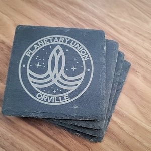 Peut inclure: Ensemble de quatre sous-verres en ardoise noire avec un motif gravé argenté du logo de l'Union Planétaire et le mot "Orville".