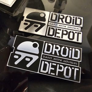 以下が含まれることがあります： 「DROID DEPOT」という文字と、スタイリッシュなドロイドの画像が描かれた、黒と銀の金属製の看板2枚。文字は未来的なフォントで、ドロイドの画像はシンプルで幾何学的なスタイルです。