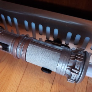 May include: A silver and brown lightsaber prop with a textured surface and a detailed handle. The lightsaber is mounted on a wall.