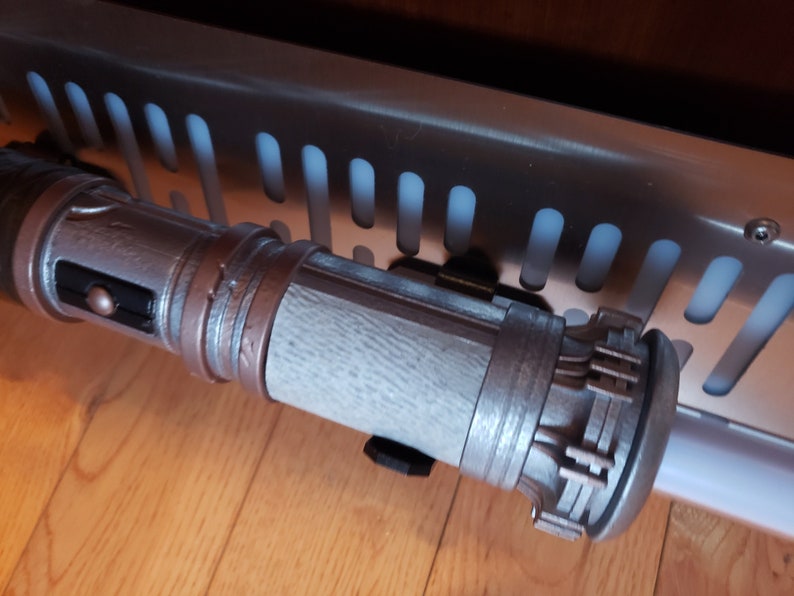 May include: A silver and brown lightsaber prop with a textured surface and a detailed handle. The lightsaber is mounted on a wall.