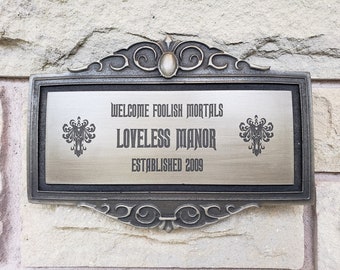 Pode incluir: Uma placa de prata com uma moldura ornamentada diz "Welcome Foolish Mortals Loveless Manor Established 2009". A placa tem dois símbolos de fantasmas pretos de cada lado do texto.