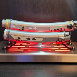 May include: Two silver lightsaber hilts with black and silver details are displayed on a metal stand with a red light. The hilts are curved and have a black diamond shape on the side.