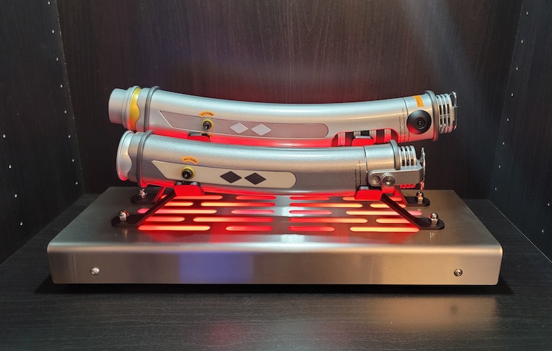 May include: Two silver lightsaber hilts with black and silver details are displayed on a metal stand with a red light. The hilts are curved and have a black diamond shape on the side.