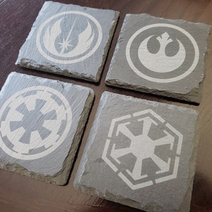 Könnte beinhalten: Vier graue Schieferuntersetzer mit weißen Star Wars-Logos. Die Logos sind das Symbol der Rebellenallianz, das Symbol des Galaktischen Imperiums, das Symbol der Ersten Ordnung und das Symbol des Jedi-Ordens.