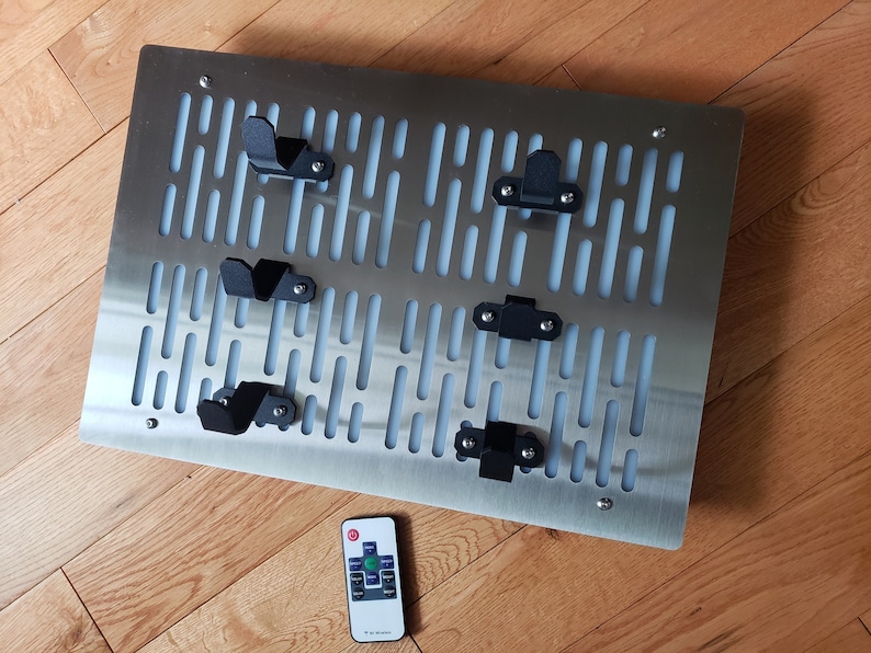 May include: A stainless steel wall-mounted rack with a grid pattern and five black metal hooks. A remote control is in the foreground.